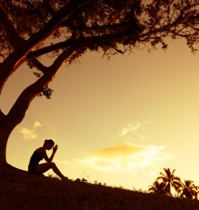 Woman sitting under tree worrying - shadowed image with sunset background