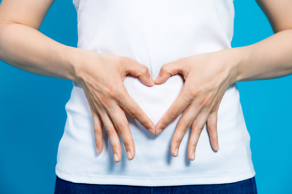 Torso of a woman in white t-shirt making the shape of a heart with her hands, over her stomach.