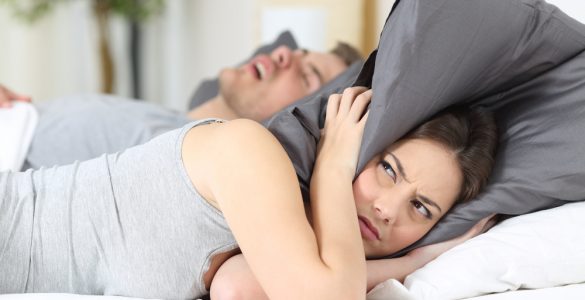 Man snoring in bed whilst woman covers her ears with a pillow