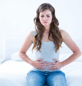 Young woman sat on a bed clutching her stomach to represent tummy ache