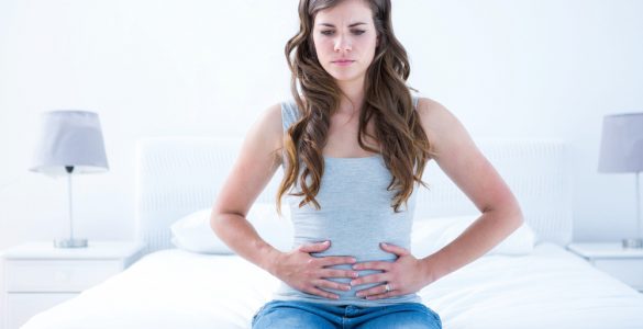 Young woman sat on a bed clutching her stomach to represent tummy ache