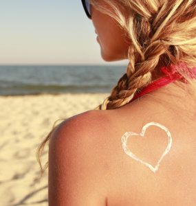 Woman on beach facing the sea with sun scream in a heart shape on her back to represent summer skin