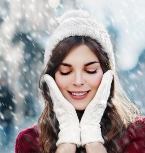 Close up of woman outdoors in the snow with her hands in gloves by her face to show winter skincare