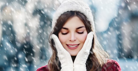 Close up of woman outdoors in the snow with her hands in gloves by her face to show winter skincare