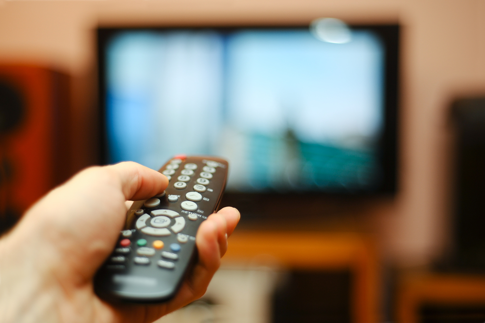 Close up of someone using a remote control with TV in the background